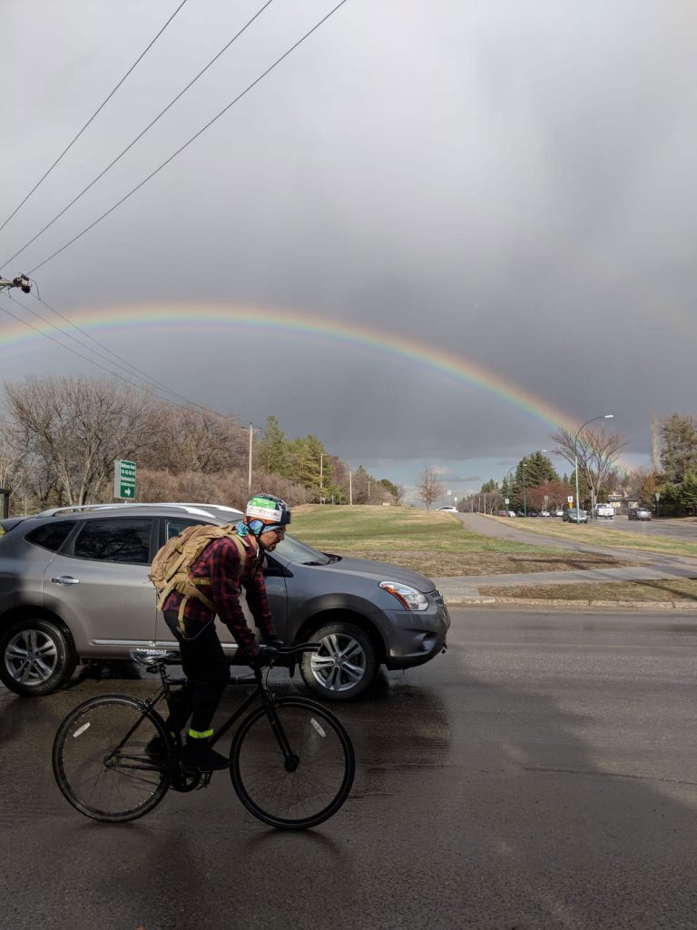 A cyclist riding in Saskatoon, a rainbow behind.
