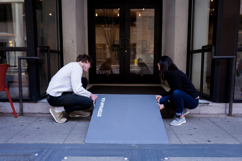 Two people crouch as they place a grey, transportable, wooden ramp in front of a business that has one step up to its front door.