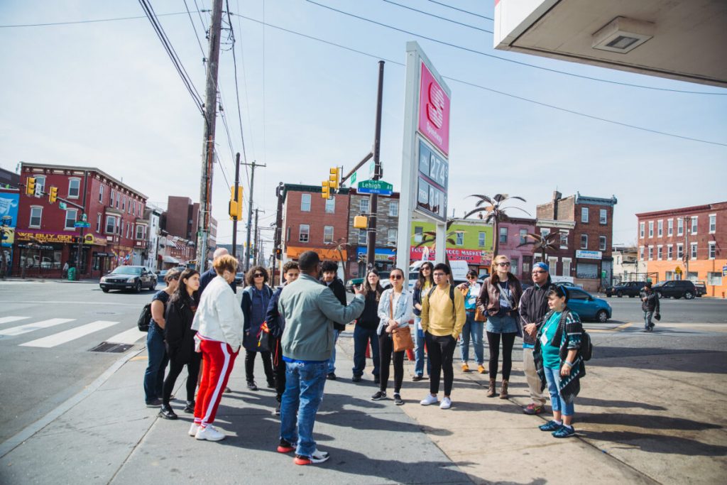 A large group of people stands in a semi-circle on a street in Philadelphia, listening to a speaker