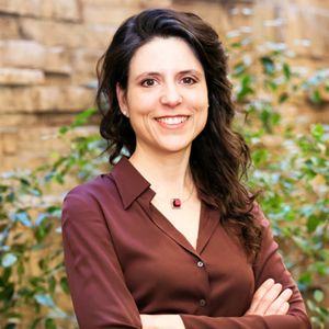 A dark-haired woman smiling warmly at the camera wearing a burgundy blouse.