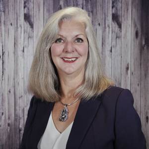 A white woman with long blond hair smiling to the camera wearing a black blazer, white shirt and a long necklace.